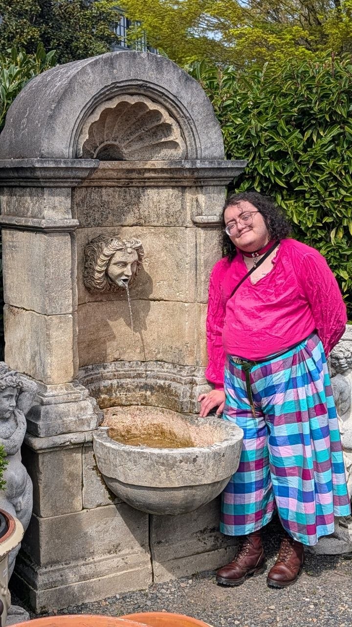 The author, a fat Latino transmasc, posing in front of an ornate wall fountain while dressed in a hot pink 18th century shirt and blue and pink checked voluminous pants - the titular slops.
